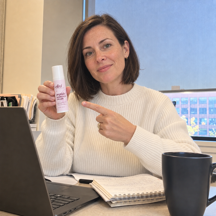 Woman in a cream sweater holding Elixi Peptide Under-Eye Cream at a desk